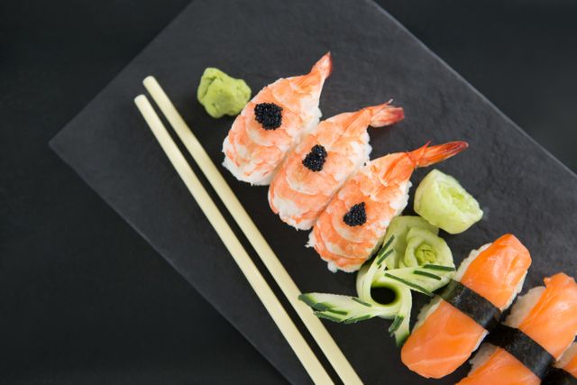 This vibrant image features beautifully arranged shrimp and salmon sushi on a sleek slate plate, accompanied by chopsticks and garnishes of wasabi and cucumber. Ideal for use in food blogs, Japanese cuisine promotions, restaurant menus, social media posts, and culinary websites highlighting gourmet seafood dishes.