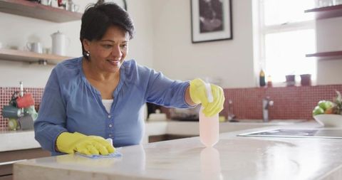 Senior Indian Woman Cleaning Kitchen Countertop Wearing Yellow Gloves and Blue Blouse