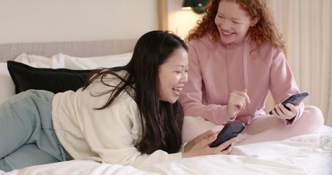 Two friends laughing and scrolling smartphones together on cozy bed in loungewear