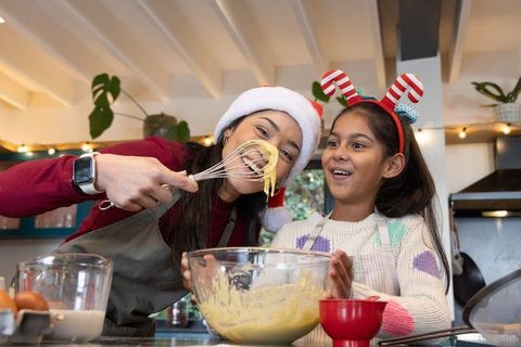 Diverse mother and daughter baking holiday treats, tasting batter, santa hat fun