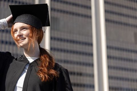 Redhead graduate smiling in black gown and mortarboard against modern city skyscraper