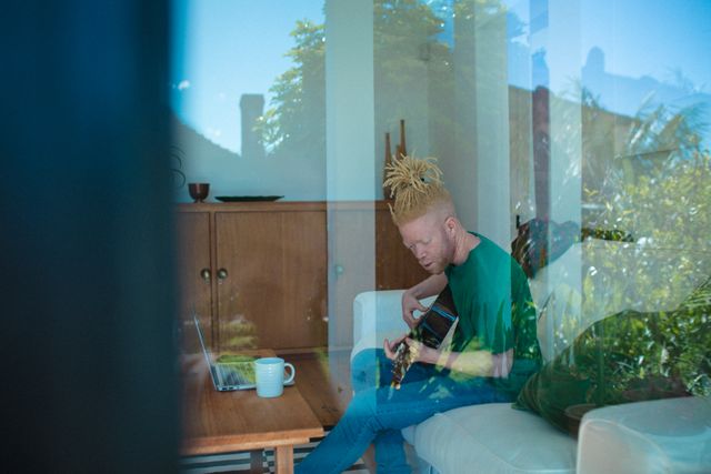 Albino African American man seen through window playing guitar in living room. Ideal for themes of leisure, relaxation, home lifestyle, music, and creativity. Suitable for articles, blogs, and advertisements focusing on home activities, personal hobbies, and indoor relaxation.