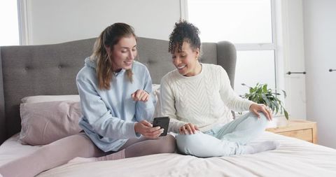 Diverse female friends sharing smartphone on bed, laughing and scrolling together