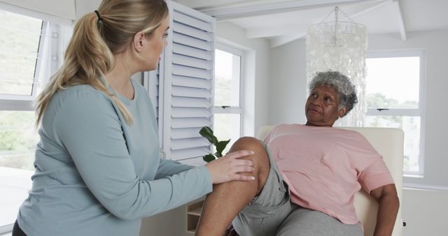 Senior woman receiving physical therapy at home from a female caregiver. Woman sitting on a chair lifting her knee while therapist supports and guides the movement. Useful for depicting home healthcare services, elderly rehabilitation, physical therapy practices, medical treatments, and senior wellness.