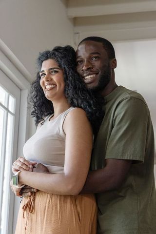 Sunlit interracial couple embracing by window, smiling partners sharing intimate moment