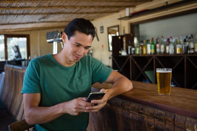 Young man in a green shirt sitting at a restaurant counter, using his phone. A glass of beer is on the counter. Ideal for use in lifestyle blogs, social media, technology, and leisure-related content.