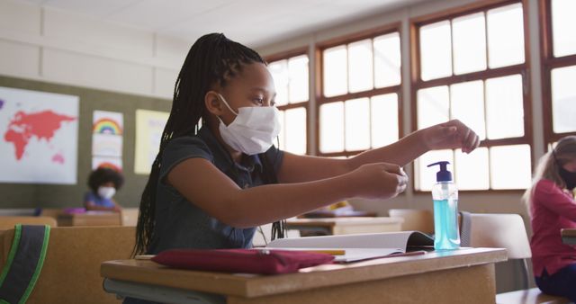 Young student uses hand sanitizer at desk wearing a mask, maintaining safety practices. Useful for content related to education during pandemic, hygiene practices in schools, child health, and COVID-19 measures in educational environments.