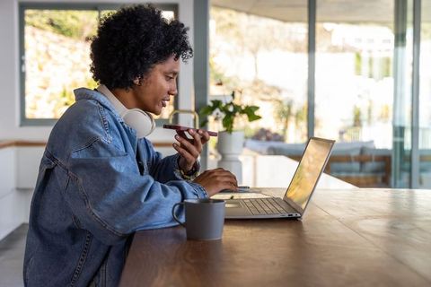 African american woman working from home on laptop speaking on smartphone with headphones