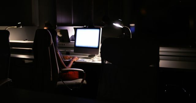 Person working in a dimly lit modern office, with light from a computer screen illuminating the dark surroundings. This image is ideal for illustrating concepts of dedication, tight deadlines, and focused work environments. Useful for articles or presentations about technology, work ethics, night shifts, and the modern work world.