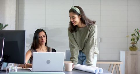 Diverse female coworkers collaborating over laptop and blueprints in creative studio office