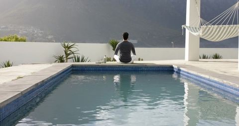 Indian man sitting cross-legged on rooftop pool deck facing mountain valley, hammock