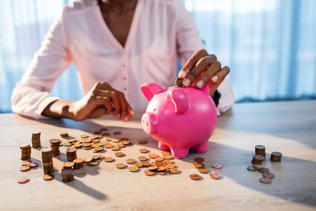 Woman placing coins into a pink piggy bank on a table, surrounded by scattered coins. Ideal for illustrating concepts of financial planning, budgeting, savings, and personal finance. Useful for articles, blogs, and advertisements related to money management, banking, and investment strategies.