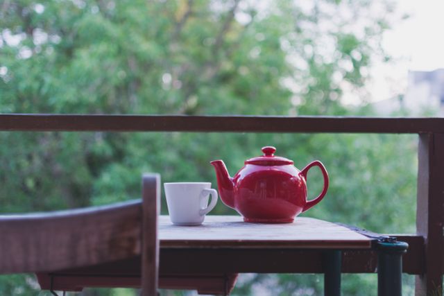 Image shows a red teapot and white mug on a wooden table with a natural green backdrop. Perfect for use in articles about morning routines, serenity, relaxation, and outdoor dining experiences.