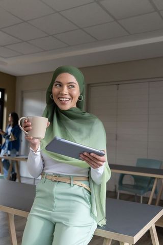 Smiling muslim businesswoman wearing green hijab holding tablet and mug in modern office