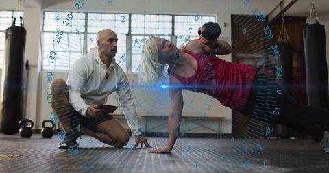Woman balancing side plank holding dumbbell while trainer recording tablet data with hud overlay