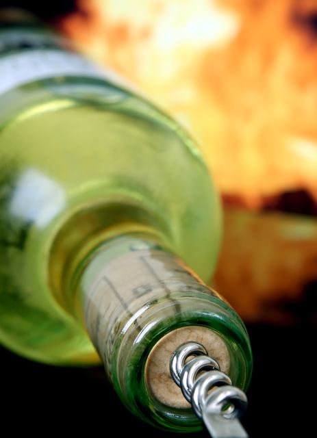 Close-up view of a white wine bottle being opened with a corkscrew against a fiery background. Ideal for advertisements and marketing related to beverages, wineries, wine tasting events, or cooking with wine. Evokes a sense of warmth, passion, and intensity.
