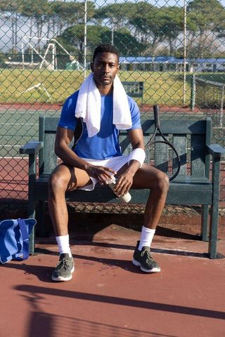 African american tennis player sitting on bench holding water bottle and racket post match