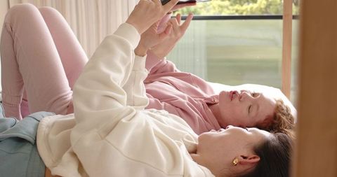 Friends lounging on bed scrolling smartphone together enjoying morning light and greenery
