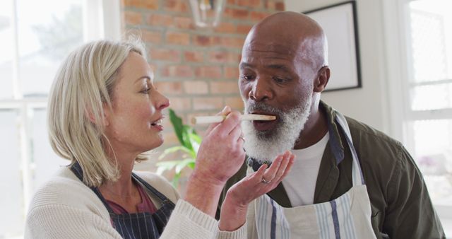 Biracial senior couple wearing aprons tasting food while cooking in the kitchen at home. retirement senior couple lifestyle living concept