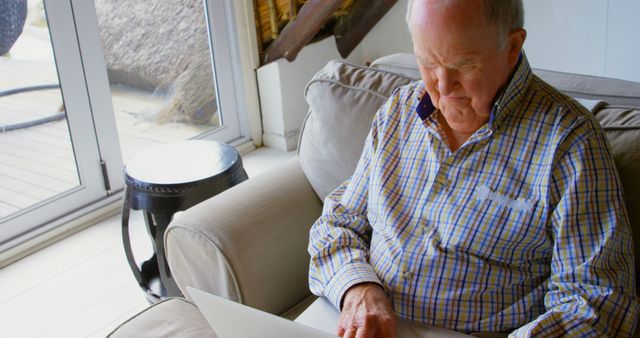 Elderly man wearing a plaid shirt sitting on sofa using laptop with focus. Bright natural light coming through glass doors in background suggesting modern and cozy interior. Suitable for use in articles, blogs, advertisements about senior citizens embracing technology, online communication, remote work, or staying connected with family.
