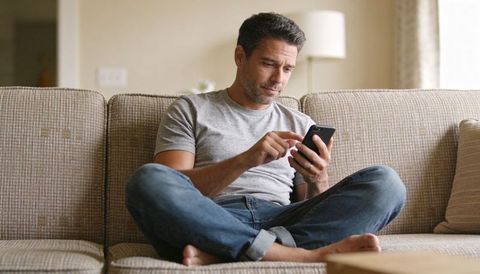 Man scrolling smartphone on beige sofa, casual home lifestyle, relaxed remote communication