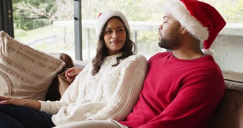 Diverse couple wearing santa hats relaxing and chatting on couch by sliding glass door