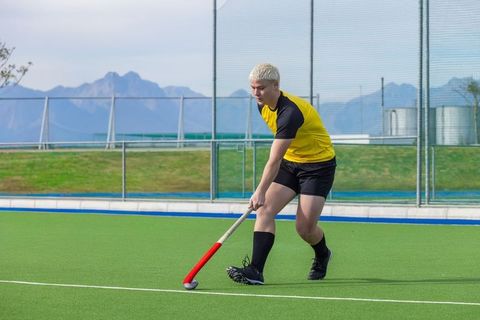 Field hockey player dribbling with red stick on green turf near mountain range