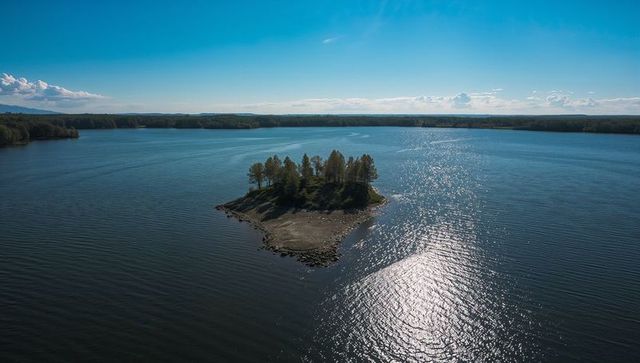 Drone capturing small tree-covered island with sun reflecting on lake water at horizon