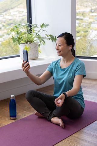 Mature asian woman practicing yoga at home holding smartphone wearing earbuds smiling