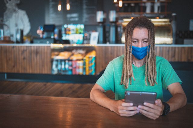 Biracial man with dreadlocks wearing a mask is sitting at a table in a modern cafe using a tablet. The cafe has a cozy and contemporary interior with a counter displaying snacks and beverages in the background. This image can be used to depict themes of technology use, urban lifestyle, small businesses, and social distancing during the pandemic.