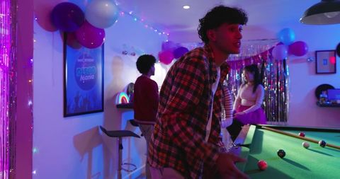 Diverse friends playing pool at neon birthday party in decorated basement with balloons