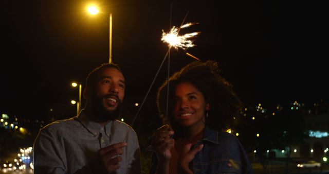 Romantic diverse couple smiling and holding sparklers in city street at night. City living, romance, love, relationship, free time and lifestyle, unaltered.