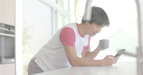 Leaning asian man holding mug and checking phone during casual morning in bright modern kitchen