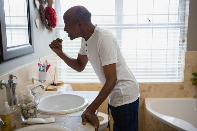 Senior man brushing teeth by sink in bathroom at home. Ideal for topics on dental hygiene, morning routines, self-care, and elderly health. Can be used in articles, blogs, and advertisements promoting dental products, health care for seniors, and personal care routines.