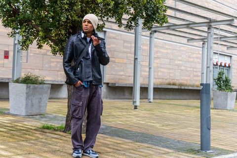 Young man standing in urban plaza holding phone to face wearing beanie and leather jacket