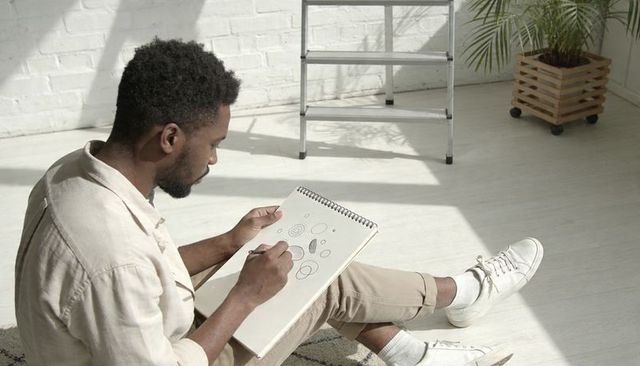 Young black artist sketching geometric shapes in sunlit minimalist studio with sketchbook