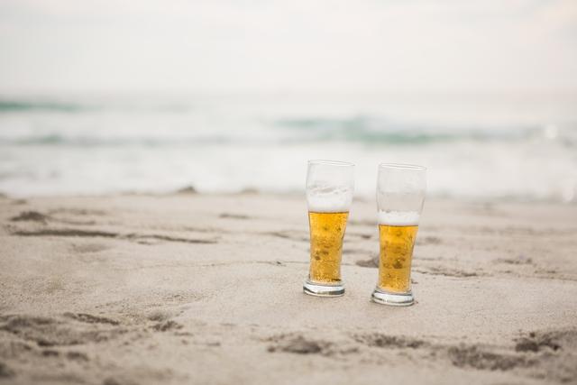 Two beer glasses are placed on sandy beach with ocean waves in background. Ideal for use in travel brochures, vacation advertisements, summer promotions, and lifestyle blogs focusing on relaxation and leisure.
