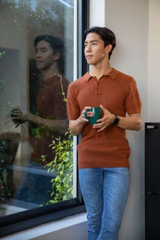Young asian man standing by window holding green mug wearing rust knit polo and smartwatch