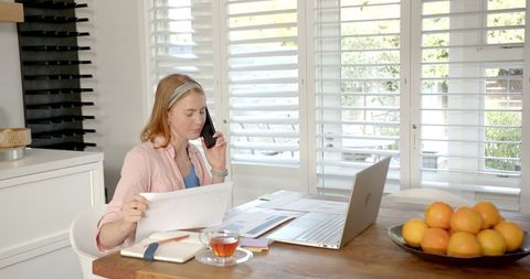 Woman Reviewing Documents at Home Office with Laptop and Smartphone