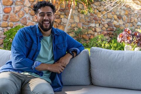 Smiling man sitting on outdoor sofa leaning back on sunny backyard patio with stonewall