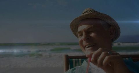 Senior man sipping cold drink on sunny beach wearing straw hat relaxing by ocean waves