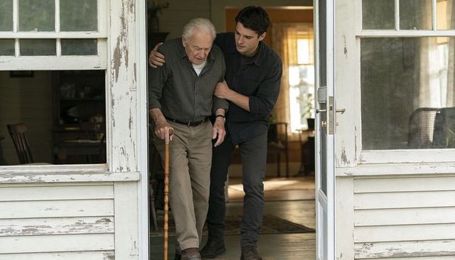 Young caregiver helping elderly man walk through porch doorway holding wooden cane