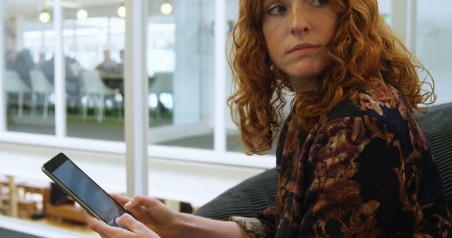 Young Caucasian woman looks at her phone in an office setting. She appears concerned or puzzled by the content on her device.