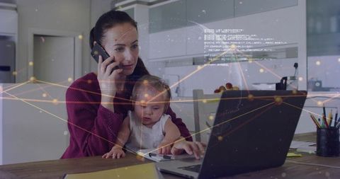 Working mom juggling baby while taking call and typing on laptop in modern kitchen