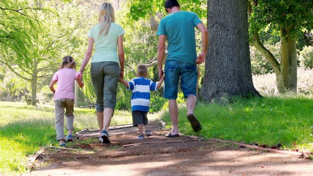 A young family, consisting of two adults and two children, enjoying a walk on a wooded path during a sunny day. Ideal for use in advertisements and campaigns related to family activities, nature exploration, parenting, leisure activities, health and wellness, and supplements connecting families with outdoor experiences.