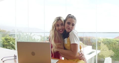 Two young women hugging and smiling on glass balcony with open laptop and coastal view