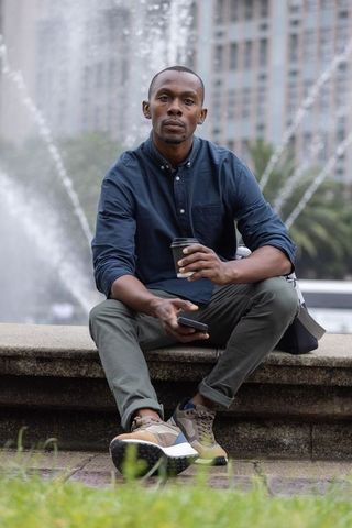 African man sitting on fountain holding coffee cup and checking phone in urban plaza