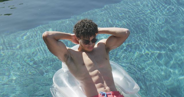 Muscular biracial man in sunglasses relaxing on inflatable in sunny swimming pool. Summer, vacations and relaxation, unaltered.