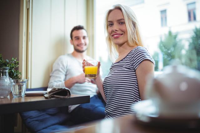 Woman smiling while holding a glass of orange juice in a cafe. Ideal for use in lifestyle blogs, social media posts, advertisements for cafes or restaurants, and articles about healthy living and socializing.