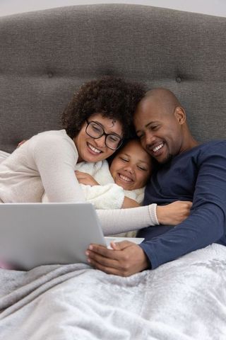 Smiling african american family cuddling in bed using laptop for cozy digital togetherness